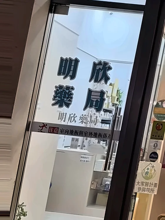 明欣藥局入口，玻璃門上印有「明欣藥局」字樣，門旁貼有大家醫計畫參與院所標示，店內可見藥品陳列櫃
