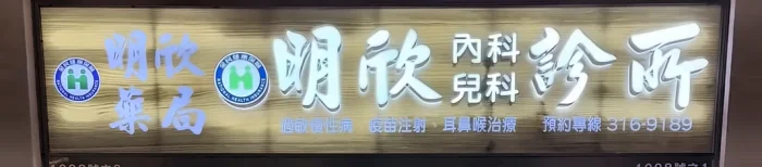 診所橫式招牌：左側為「明欣藥局」與健保特約標章，右側為「明欣 內科 兒科 診所」，下排小字寫著過敏慢性病、疫苗注射、耳鼻喉治療與預約專線 316-9189