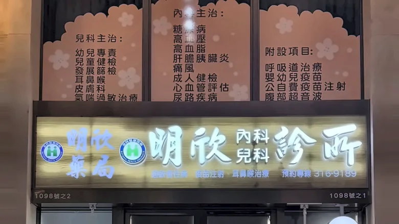 夜間的明欣診所店面：上方橘色看板列出兒科主治、內科主治與附設項目，下方燈箱寫著明欣藥局與明欣內科兒科診所