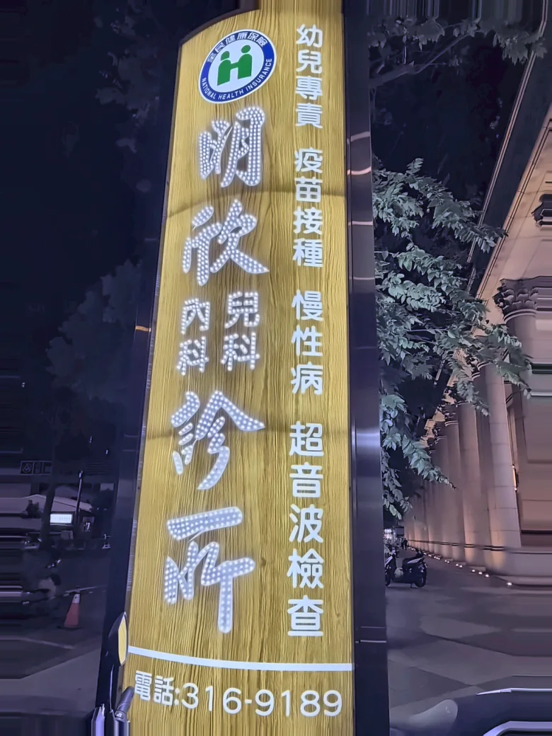 騎樓柱上的直式燈箱夜間亮著，寫著幼兒專責、疫苗接種、慢性病、超音波檢查，下方標示電話 316-9189，兩側可見騎樓廊柱與人行道